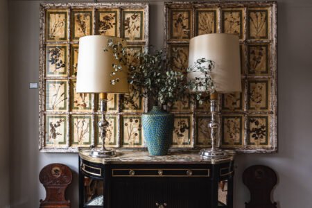 Antique mirrored panels with botanical patterns displayed behind a vintage console table and decorative lamps, by D&E Decor.